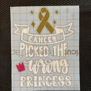 May include: A white vinyl decal with the text "CANCER PICKED THE WRONG PRINCESS". The design includes a gold ribbon, a pink crown, and star accents. The decal is on a light blue grid background.