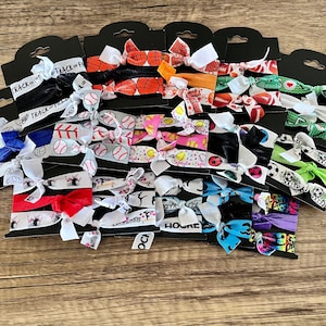 May include: A variety of colorful hair ties with different sports themes, including soccer, basketball, volleyball, softball, track, cheer, and hockey. The hair ties are arranged in rows on black cards.