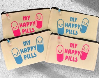 May include: Four canvas zipper pouches with the text "MY HAPPY PILLS" and a smiling pill graphic in pink and blue. The pouches have a black zipper and a small charm. The phrase "Merry for a Cause" is at the bottom.