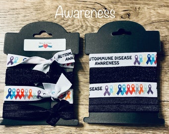 May include: Two sets of black hair ties with white ribbon and colorful awareness ribbons. The ribbon reads "Autoimmune Disease Awareness". The packaging is labeled "Marry for a Cause".