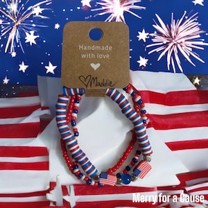 May include: Three patriotic bracelets with red, white, and blue beads and flag charms. The bracelets are displayed on a white surface with an American flag background. A tag reads "Handmade with love" and "Merry for a Cause".