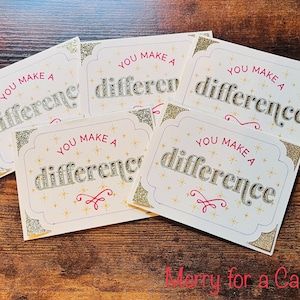 Puede incluir: Cinco tarjetas blancas con bordes de purpurina dorada y estrellas doradas. Cada tarjeta tiene el texto "You Make A Difference" en letras plateadas con acentos rosados. El texto "Merry for a Cause" está escrito en rosa en la parte inferior de la imagen.