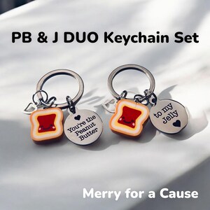 May include: A set of two keychains featuring a peanut butter and jelly sandwich theme. One keychain has a charm with the text "You're the Peanut Butter" and the other keychain has a charm with the text "to my Jelly".