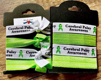 May include: Three green and white elastic hair ties with the text "Cerebral Palsy Awareness" and a green ribbon. The hair ties are packaged on a white card with the text "Merry for a Cause".