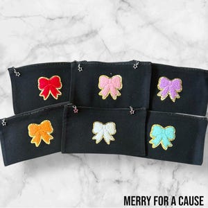 Könnte beinhalten: Sechs schwarze Reißverschlusstaschen mit verschiedenen farbigen Schleifen, die auf der Vorderseite gestickt sind. Die Schleifen sind rosa, rot, lila, orange, weiß und blau. Die Taschen haben einen Sternanhänger am Reißverschluss. Der Text "MERRY FOR A CAUSE" ist unten im Bild gedruckt.