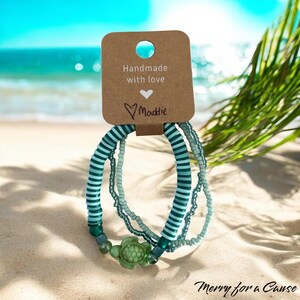 May include: Three beaded bracelets in shades of teal and white, with a turtle charm. The bracelets are displayed on a sandy beach with a tag that reads "Handmade with love" and "Maddie". The background shows the ocean and palm leaves.