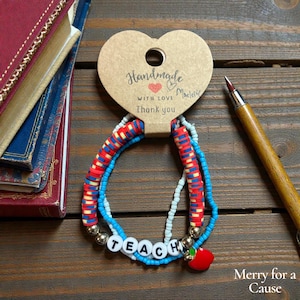 May include: A handmade bracelet with red, white, and blue beads, spelling "TEACH" with white letter beads. It includes a red apple charm and a heart-shaped tag that reads "Handmade with love".
