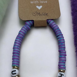 May include: Purple and blue striped beaded bracelet with white beads spelling out the word "UR ENOUGH". The bracelet has a tag that says "Handmade with love Maddie".