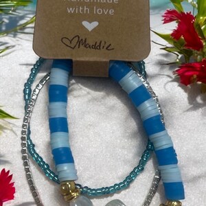 May include: Set of three bracelets with blue and white striped beads, turquoise beads, and silver chain with a white seashell charm. The bracelets are handmade with love by Maddie. The tag also says "Merry for a cause".