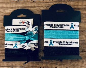 May include: Two sets of hair ties with the text "Fragile X Syndrome Awareness" and a blue ribbon. The hair ties are packaged in clear plastic with a black header card that says "Merry for a Cause".