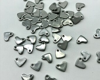 Könnte beinhalten: Eine große Menge silberfarbener, herzförmiger Metall-Anhänger mit einer kleinen Schlaufe oben. Die Anhänger sind auf einer weißen Oberfläche verstreut.