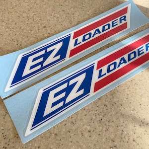 May include: Two blue and white "EZ Loader" decals on a light blue background. The decals have red stripes and white lettering.