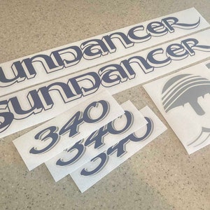 Puede incluir: Un conjunto de calcomanías de vinilo blanco con letras azules. Las calcomanías incluyen las palabras "Sundancer" y "340".