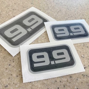 May include: Three stickers with the number "9.9" in a gray, rectangular design with a black border. The stickers are on a white background and are arranged on a speckled, light brown surface.
