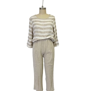 Puede incluir: Una blusa de lino a rayas con un corte holgado y mangas 3/4 combinada con pantalones de lino a juego. La blusa y los pantalones son de color beige claro con rayas blancas.