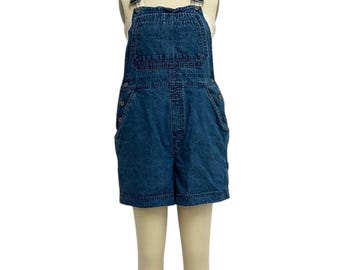 Puede incluir: Pantalones cortos de peto de mezclilla azul con tirantes ajustables, un bolsillo de peto y cierres laterales con botones. Los pantalones cortos se exhiben en un maniquí. La prenda tiene un estilo clásico e informal.