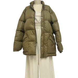 Può includere: Giacca puffer verde oliva con collo alto, cerniera frontale e chiusure a scatto. La giacca ha due tasche frontali e un design trapuntato. La giacca è esposta su un manichino con un top e pantaloni color crema.
