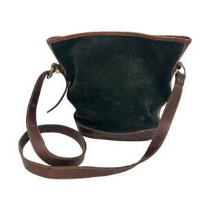 Peut inclure: Sac à bandoulière en daim vert foncé avec garniture et bandoulière en cuir marron. Le sac a une forme de seau avec un fond arrondi et une longue bandoulière réglable. La bandoulière en cuir est cousue avec du fil blanc.