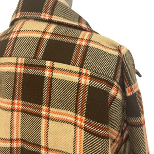 May include: Close-up of a brown, tan, and orange plaid jacket. The jacket has a classic plaid pattern with a collar and a button-down front. The fabric appears to be a woven material, and the overall style is vintage.