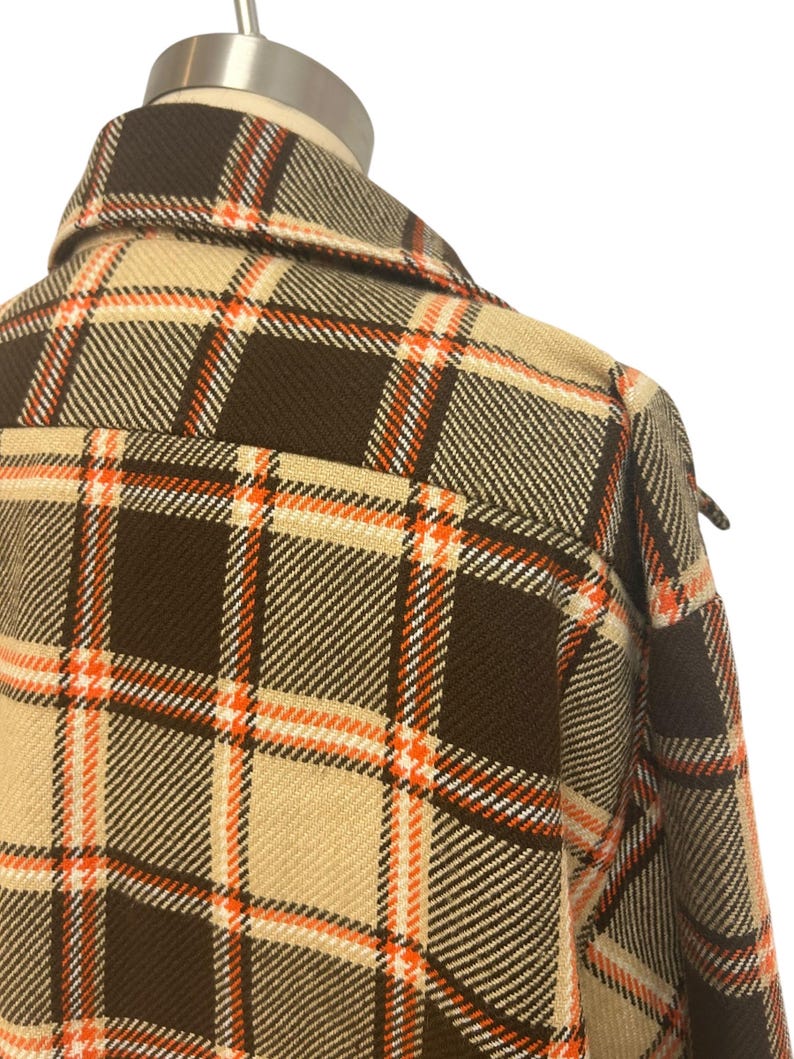 May include: Close-up of a brown, tan, and orange plaid jacket. The jacket has a classic plaid pattern with a collar and a button-down front. The fabric appears to be a woven material, and the overall style is vintage.