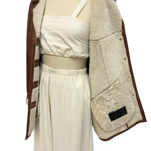 May include: Brown leather jacket with a cream-colored shearling lining. The jacket has a classic design with a button closure and a warm, inviting look. The jacket is displayed on a mannequin.