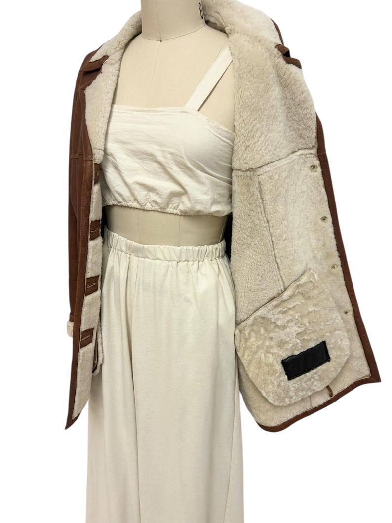 May include: Brown leather jacket with a cream-colored shearling lining. The jacket has a classic design with a button closure and a warm, inviting look. The jacket is displayed on a mannequin.