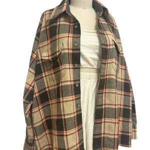 May include: A tan, brown, and orange plaid flannel shirt with two chest pockets and a button-down front. The shirt is displayed on a mannequin, revealing a white crop top and shorts underneath. The shirt has a classic collar and long sleeves.
