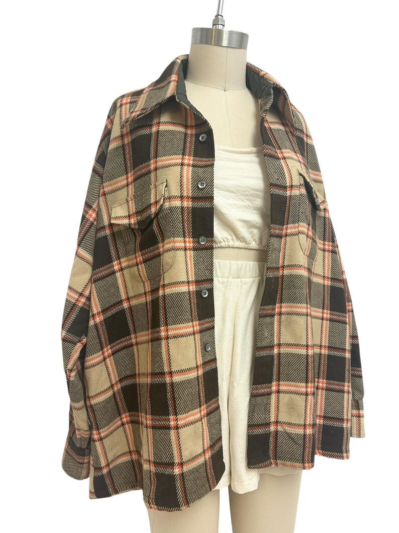 May include: A tan, brown, and orange plaid flannel shirt with two chest pockets and a button-down front. The shirt is displayed on a mannequin, revealing a white crop top and shorts underneath. The shirt has a classic collar and long sleeves.
