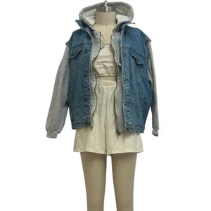 May include: A denim jacket with an attached grey hooded sweatshirt, worn with a cream crop top and shorts. The jacket has a button-up front and pockets. The shorts have an elasticated waistband.