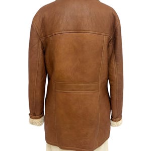May include: A brown leather jacket with a cream-colored shearling collar and cuffs. The jacket has a classic design with a button closure and a tailored fit. The back of the jacket features a horizontal seam across the upper back.