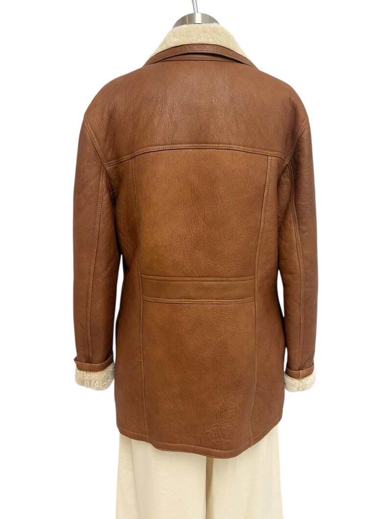 May include: A brown leather jacket with a cream-colored shearling collar and cuffs. The jacket has a classic design with a button closure and a tailored fit. The back of the jacket features a horizontal seam across the upper back.
