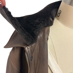 Puede incluir: Primer plano de una chaqueta de cuero marr&oacute;n con un cuello de piel marr&oacute;n oscuro. La chaqueta tiene una cremallera y un cierre de bot&oacute;n. El cuello se levanta para mostrar el forro suave y peludo.