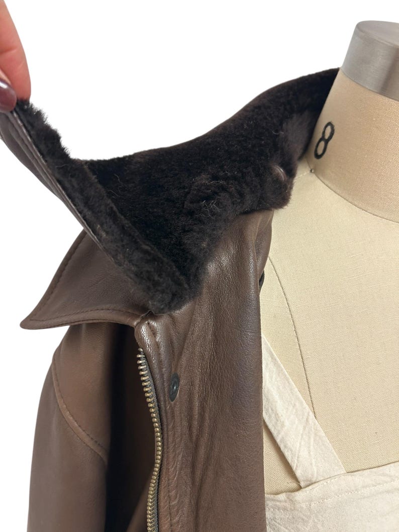Puede incluir: Primer plano de una chaqueta de cuero marr&oacute;n con un cuello de piel marr&oacute;n oscuro. La chaqueta tiene una cremallera y un cierre de bot&oacute;n. El cuello se levanta para mostrar el forro suave y peludo.