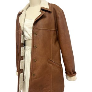 May include: A brown leather jacket with a cream-colored shearling lining and collar. The jacket has a button-up front, two front pockets, and shearling cuffs. The jacket is displayed on a mannequin.