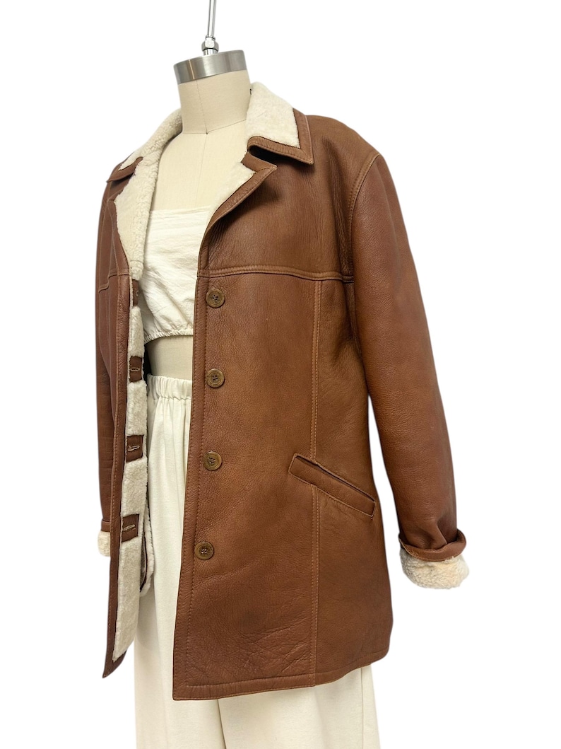 May include: A brown leather jacket with a cream-colored shearling lining and collar. The jacket has a button-up front, two front pockets, and shearling cuffs. The jacket is displayed on a mannequin.