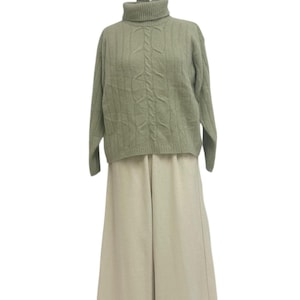 Könnte beinhalten: Ein salbeigrüner Rollkragenpullover mit Zopfmuster, kombiniert mit cremefarbenen Hosen mit weitem Bein. Der Pullover hat lange Ärmel und einen gerippten Rollkragen. Die Hose ist hoch tailliert und hat eine fließende Silhouette. Das Outfit wird vor weißem Hintergrund präsentiert.