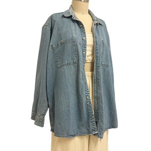 Op de afbeelding: Een lichtblauw spijkeroverhemd met twee borstzakken en knoopsluitingen. Het shirt met lange mouwen is open en onthult een cr&egrave;mekleurige top en een beige broek. Het shirt wordt op een mannequin getoond.