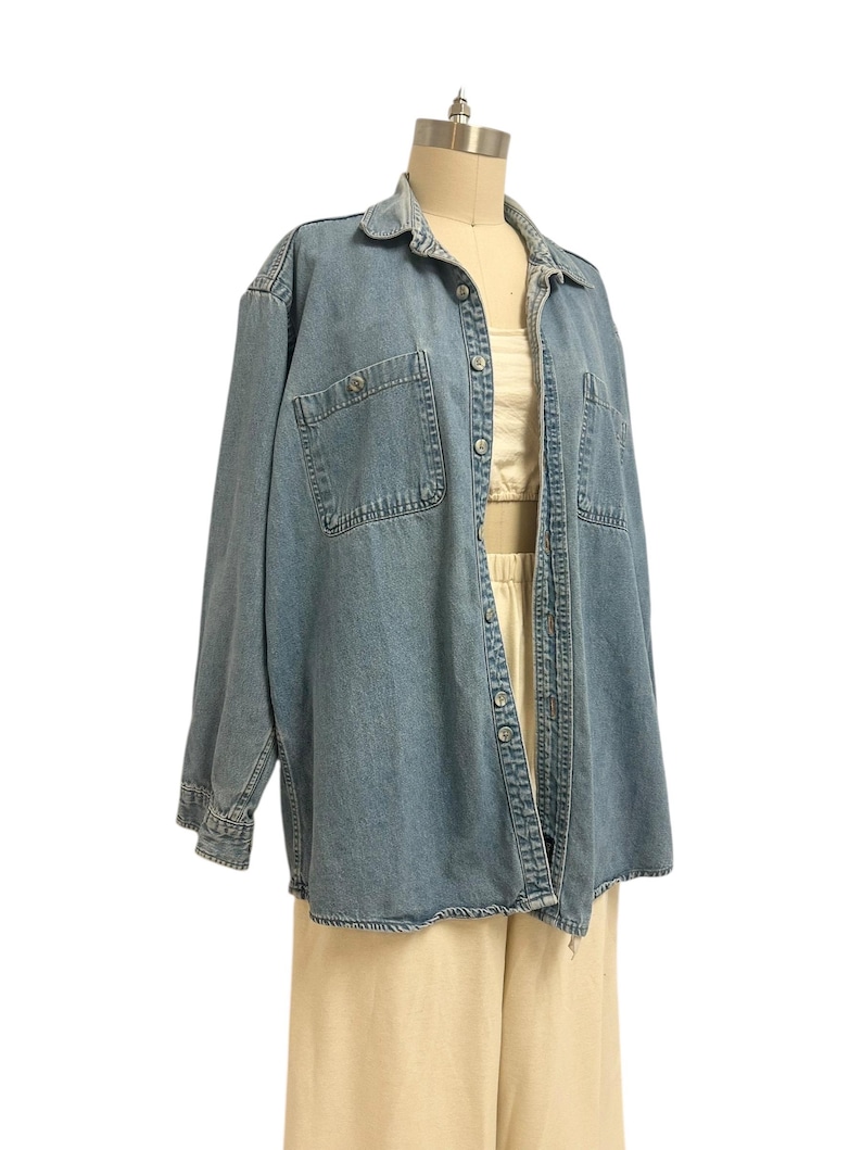 Op de afbeelding: Een lichtblauw spijkeroverhemd met twee borstzakken en knoopsluitingen. Het shirt met lange mouwen is open en onthult een cr&egrave;mekleurige top en een beige broek. Het shirt wordt op een mannequin getoond.