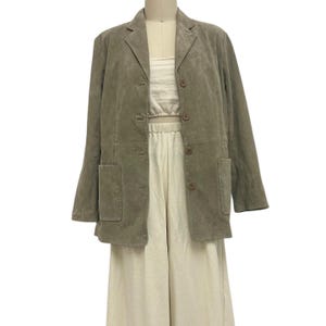 Puede incluir: Una blazer de ante verde salvia con solapa de muesca, tres botones y dos bolsillos delanteros. La blazer se lleva sobre una camiseta y un pantalón de color crema. El conjunto se exhibe en un maniquí.