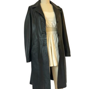 May include: A long, black leather coat with a classic lapel collar and button closure. The coat is displayed on a mannequin, revealing a cream-colored top and skirt underneath. The coat appears to be knee-length.