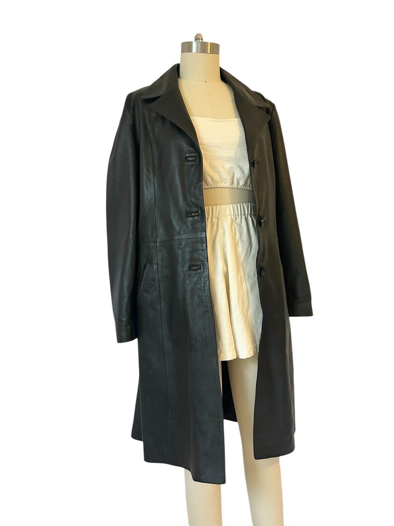 May include: A long, black leather coat with a classic lapel collar and button closure. The coat is displayed on a mannequin, revealing a cream-colored top and skirt underneath. The coat appears to be knee-length.