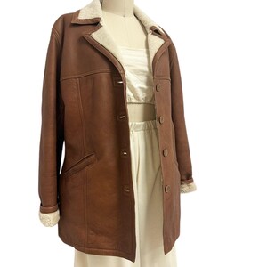 May include: A brown leather coat with a cream-colored shearling collar and cuffs. The coat has a button-up front, two front pockets, and is displayed on a mannequin. The coat is a classic style.