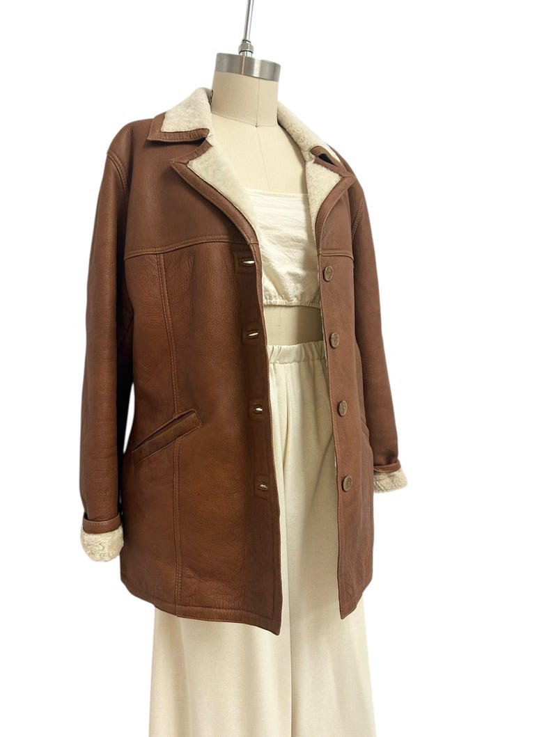 May include: A brown leather coat with a cream-colored shearling collar and cuffs. The coat has a button-up front, two front pockets, and is displayed on a mannequin. The coat is a classic style.