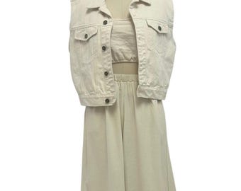 Könnte beinhalten: Ein dreiteiliges beiges Outfit, das auf einer Schaufensterpuppe präsentiert wird. Das Ensemble umfasst eine ärmellose Jeansjacke mit Knopfverschlüssen und zwei Vordertaschen, ein passendes Crop-Top und eine Hose mit weitem Bein. Das Outfit ist aus einem hellfarbigen Stoff gefertigt.