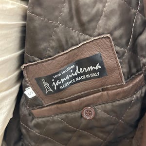 Puede incluir: Primer plano del interior de una chaqueta de cuero marr&oacute;n. El forro acolchado es visible, junto con un bolsillo de cuero marr&oacute;n y un bot&oacute;n. Una etiqueta dice "real leather janniderma FLORENCE MADE IN ITALY."