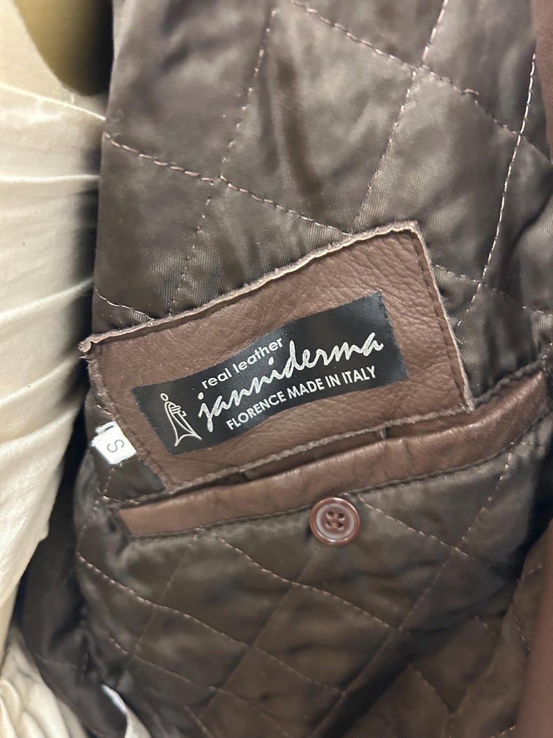 Puede incluir: Primer plano del interior de una chaqueta de cuero marr&oacute;n. El forro acolchado es visible, junto con un bolsillo de cuero marr&oacute;n y un bot&oacute;n. Una etiqueta dice "real leather janniderma FLORENCE MADE IN ITALY."