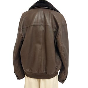 Puede incluir: Chaqueta de cuero marr&oacute;n con cuello de piel negra. La chaqueta tiene dobladillo y pu&ntilde;os acanalados. La parte trasera de la chaqueta es visible, mostrando las costuras y los detalles del panel. La chaqueta se exhibe en un maniqu&iacute;.
