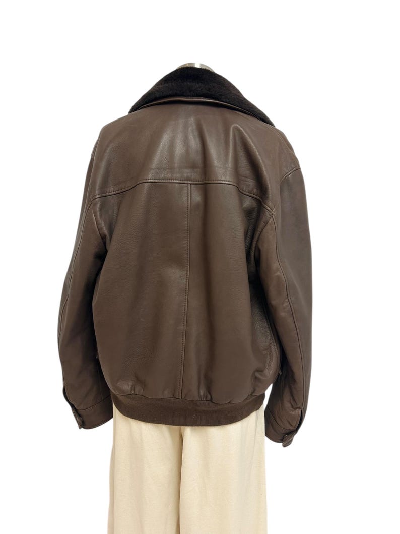Puede incluir: Chaqueta de cuero marr&oacute;n con cuello de piel negra. La chaqueta tiene dobladillo y pu&ntilde;os acanalados. La parte trasera de la chaqueta es visible, mostrando las costuras y los detalles del panel. La chaqueta se exhibe en un maniqu&iacute;.