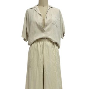 May include: A two-piece linen outfit in a neutral beige color. The top is a short-sleeved button-down shirt with a chest pocket. The bottoms are wide-legged pants with an elastic waistband. The outfit is displayed on a mannequin.
