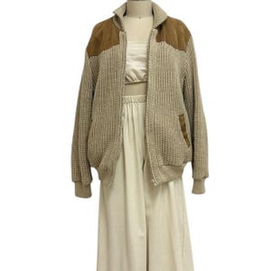 May include: A tan knit zip-up jacket with brown suede patches on the shoulders and pockets. The jacket is displayed on a mannequin with a cream-colored top and pants. The jacket has a ribbed collar and cuffs.